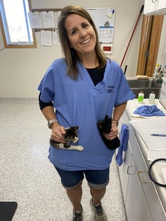 A woman in blue scrubs stands in a veterinary clinic, smiling and holding two small kittens, one in each pocket of her shirt.